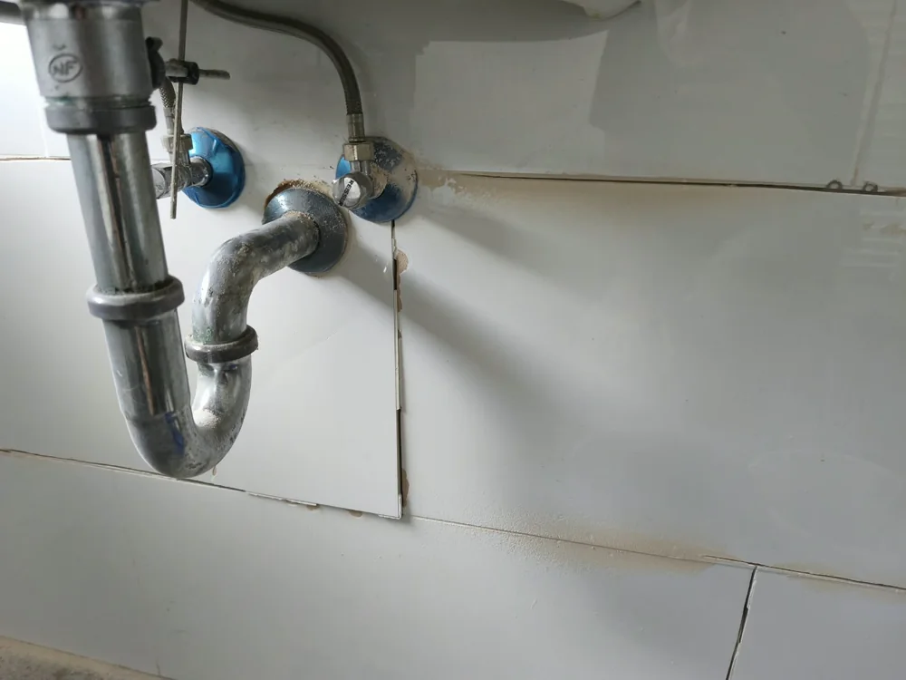 Beschädigte und offene Fliesenanschlüsse unter einem Waschtisch vor der Reparatur.